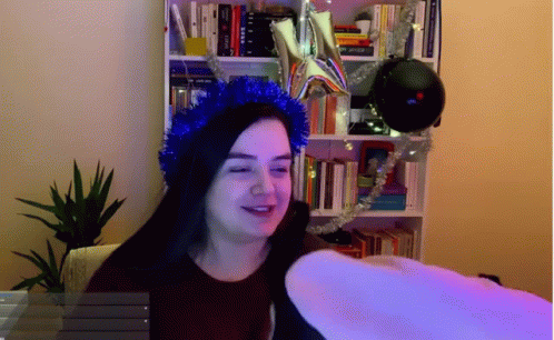 A Woman Wearing A Blue Tinsel Headband Stands In Front Of A Bookcase GIF