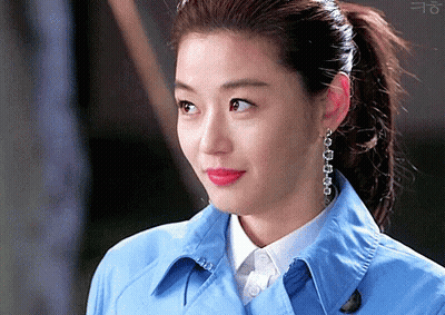 A Woman Wearing A Blue Trench Coat And Earrings Looks At The Camera GIF
