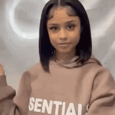 A Woman Wearing A Brown Hoodie Is Standing In Front Of A White Wall . GIF