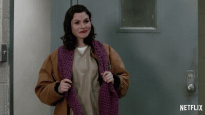 A Woman Wearing A Brown Jacket And A Purple Scarf Stands In Front Of A Netflix Logo GIF