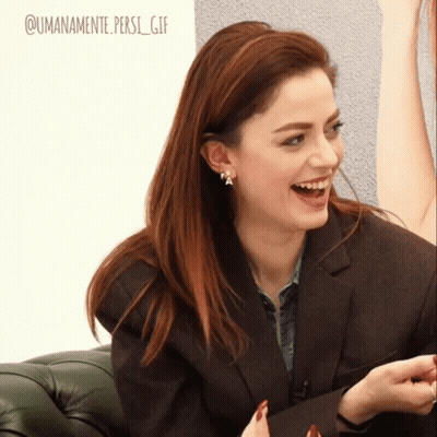 A Woman Wearing A Brown Jacket And Earrings Is Laughing With The Words @Umanamente Persi_gif Below Her GIF