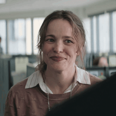 A Woman Wearing A Brown Sweater And A White Shirt Smiles For The Camera GIF