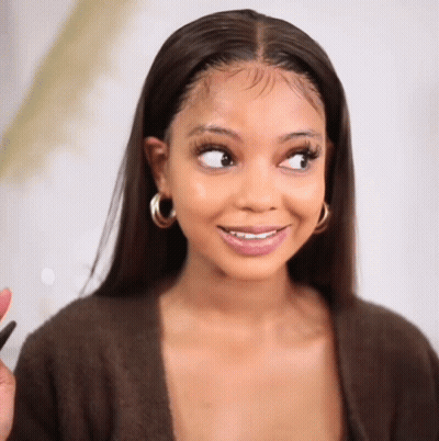 A Woman Wearing A Brown Sweater And Hoop Earrings Is Smiling And Looking To The Side . GIF