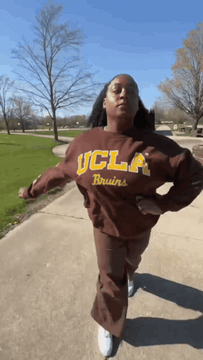 A Woman Wearing A Brown Ucla Bruins Sweatshirt Is Walking Down A Sidewalk . GIF