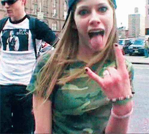 A Woman Wearing A Camouflage Shirt Is Sticking Her Tongue Out And Making A Rock On Sign GIF