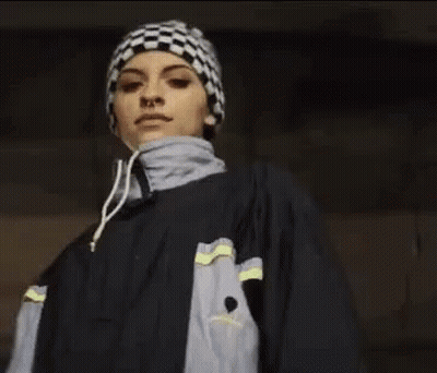A Woman Wearing A Checkered Hat And A Jacket Is Pointing At The Camera . GIF