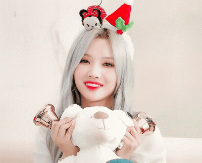 A Woman Wearing A Christmas Hat Holds A Bell In Front Of Her Ear While Holding A Teddy Bear GIF