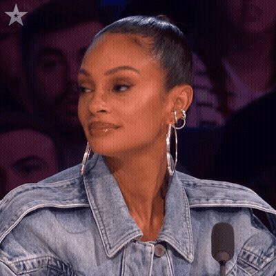 A Woman Wearing A Denim Jacket And Hoop Earrings Looks At The Camera GIF