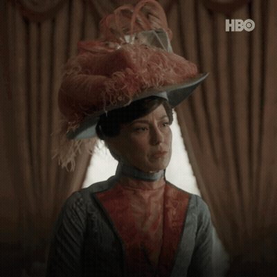 A Woman Wearing A Feathered Hat Says " What Can I Do " In White Letters GIF