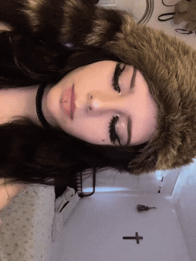 A Woman Wearing A Fur Hat And A Choker Has A Cross On The Wall Behind Her GIF