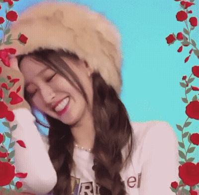 A Woman Wearing A Fur Hat And A White Shirt Is Smiling In Front Of Red Roses . GIF