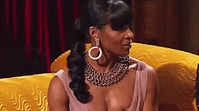 A Woman Wearing A Gold Necklace And Earrings Is Sitting On A Yellow Couch . GIF