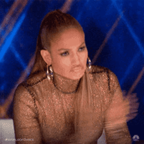 A Woman Wearing A Gold Top And Earrings Is Sitting In Front Of A Blue Background . GIF