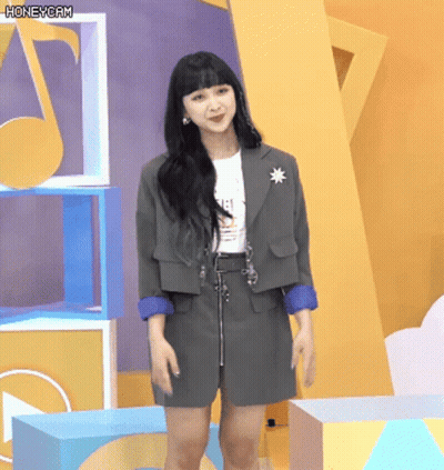 A Woman Wearing A Gray Jacket And Skirt Stands In Front Of A Yellow Wall With A Honeycam Watermark GIF