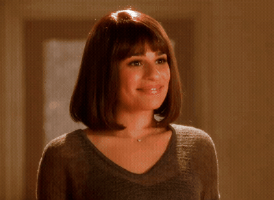 A Woman Wearing A Gray Sweater And A Necklace Smiles GIF