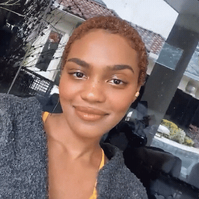 A Woman Wearing A Gray Sweater And A Yellow Top Smiles For The Camera GIF