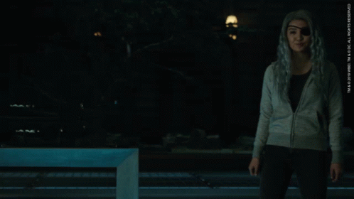 A Woman Wearing A Gray Sweater And Black Pants Stands In A Dark Room GIF