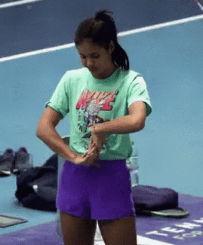 A Woman Wearing A Green Nike T-Shirt And Purple Shorts Is Standing On A Tennis Court . GIF