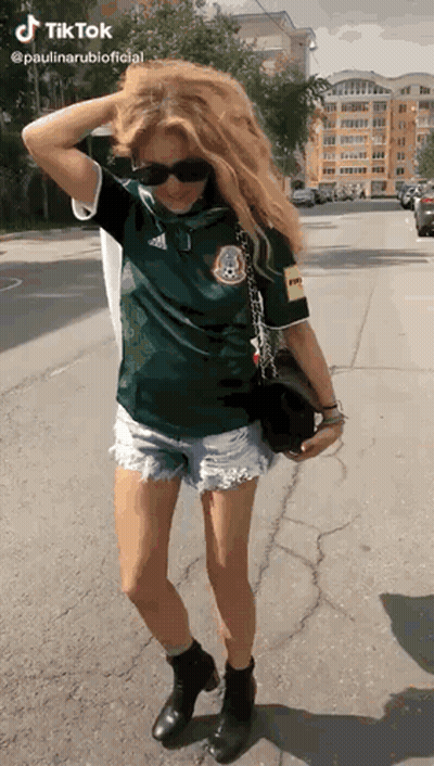 A Woman Wearing A Green Shirt And Shorts Is Standing On The Sidewalk . GIF