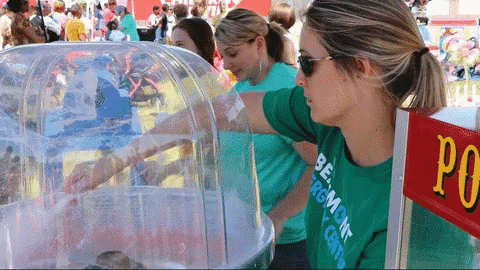 A Woman Wearing A Green Shirt That Says Burlington On It Stands In Front Of A Cotton Candy Machine GIF