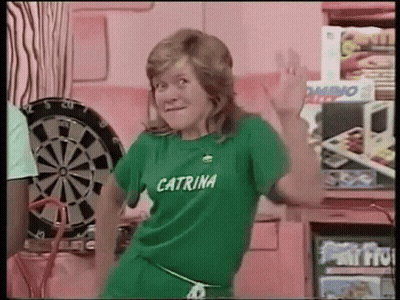 A Woman Wearing A Green Shirt That Says Caterina Is Dancing In Front Of A Dartboard . GIF