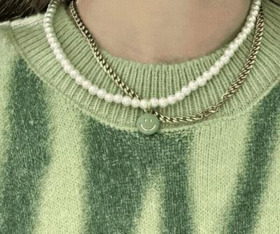 A Woman Wearing A Green Sweater And Pearl Necklaces With A Smiley Face Pendant GIF