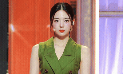 A Woman Wearing A Green Vest And Red Lipstick Is Standing In Front Of A Microphone . GIF