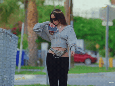 A Woman Wearing A Grey Hoodie And Black Pants Holds A Cell Phone In Her Hand GIF