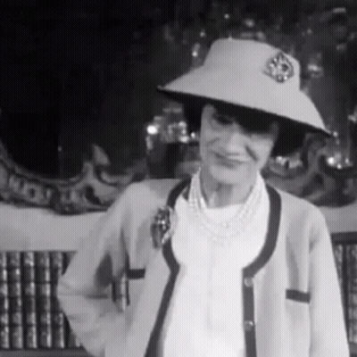 A Woman Wearing A Hat And A Cardigan Is Standing In Front Of A Bookshelf . GIF