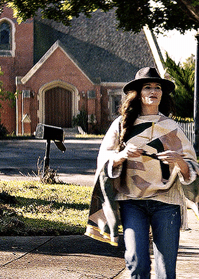 A Woman Wearing A Hat And A Poncho Is Walking Down The Sidewalk GIF