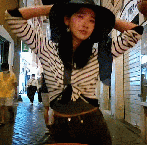 A Woman Wearing A Hat And A Striped Shirt Stands In Front Of A Building That Says ' Real Muram ' On It GIF