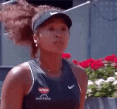 A Woman Wearing A Hat And A Tank Top Is Standing In Front Of Flowers . GIF