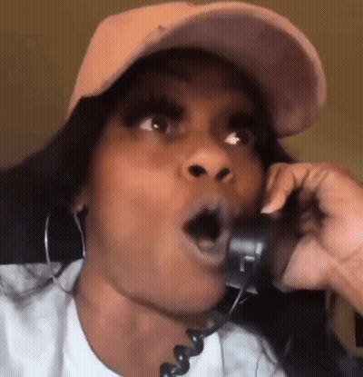 A Woman Wearing A Hat And Earrings Is Talking On A Phone With Her Mouth Open . GIF