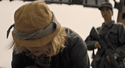 A Woman Wearing A Hat Looks Down At Something While A Man Holding A Gun Stands Behind Her GIF