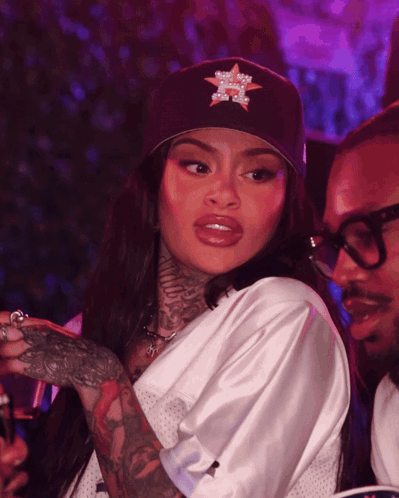 A Woman Wearing A Hat With A Star On It Stands Next To A Man Wearing Glasses GIF