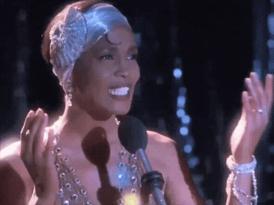 A Woman Wearing A Headband And Pearls Is Singing Into A Microphone . GIF
