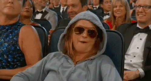 A Woman Wearing A Hoodie And Sunglasses Is Sitting In A Crowd Of People At A Movie Theater . GIF