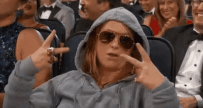 A Woman Wearing A Hoodie And Sunglasses Is Sitting In A Crowd Of People Making A Peace Sign . GIF