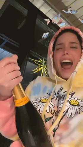 A Woman Wearing A Hoodie With Daisies On It Is Opening A Bottle Of Champagne GIF