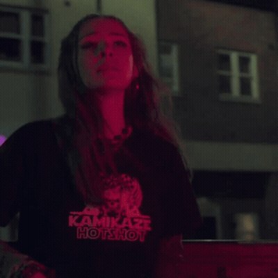 A Woman Wearing A Kamikaze Hotshot T-Shirt Stands In Front Of A Building GIF
