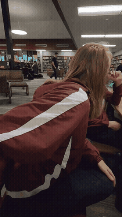 A Woman Wearing A Maroon Jacket With White Stripes On The Back Sits In A Library GIF