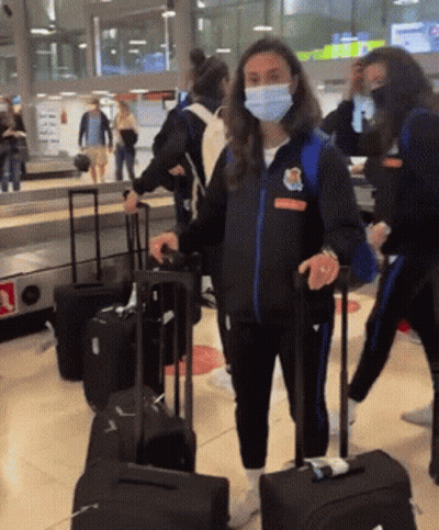 A Woman Wearing A Mask And A Jacket That Says ' Athletic ' On It Is Pulling A Suitcase GIF