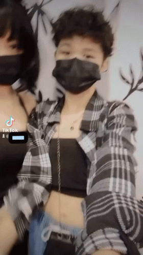 A Woman Wearing A Mask And A Plaid Shirt Is Standing Next To Another Woman GIF