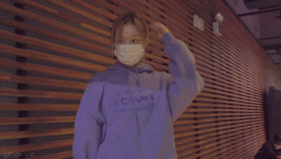 A Woman Wearing A Mask And A Purple Hoodie Is Standing In Front Of A Wooden Wall . GIF