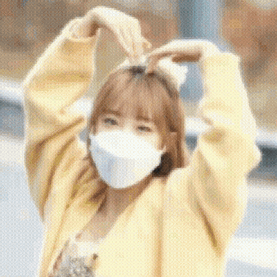 a woman wearing a mask and a yellow sweater is making a heart shape with her hands .