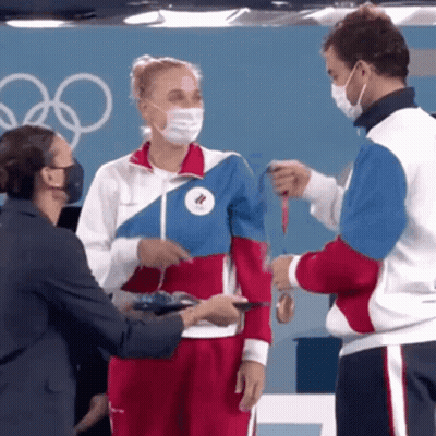 A Woman Wearing A Mask Is Being Handed A Medal By Two Men GIF