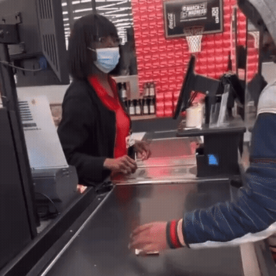 A Woman Wearing A Mask Is Standing At A Cash Register In Front Of A Sign That Says Coca Cola GIF