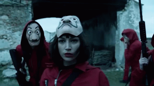 A Woman Wearing A Mask Is Standing In Front Of A Group Of People Wearing Red Jumpsuits And Holding Guns . GIF