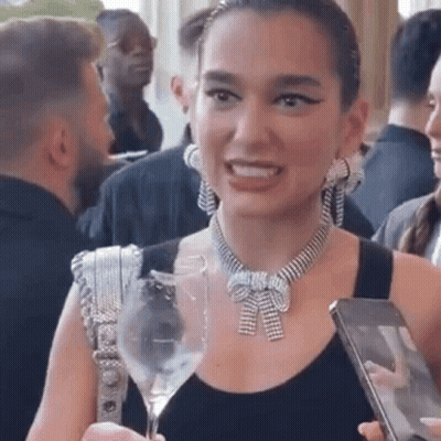 A Woman Wearing A Necklace And Earrings Is Holding A Wine Glass And A Cell Phone . GIF