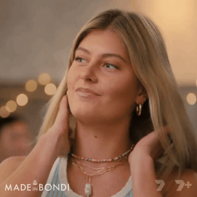 A Woman Wearing A Necklace And Earrings Is Smiling And Touching Her Hair GIF